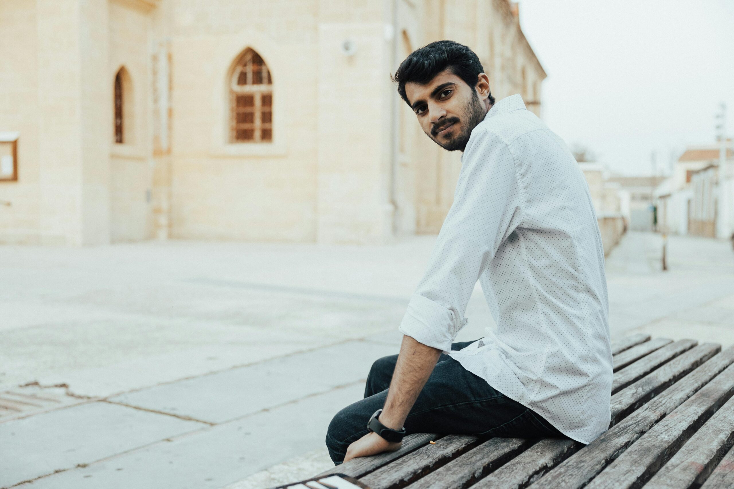 A man in white long sleeves sitting outdoors on a wooden bench, looking sideways.